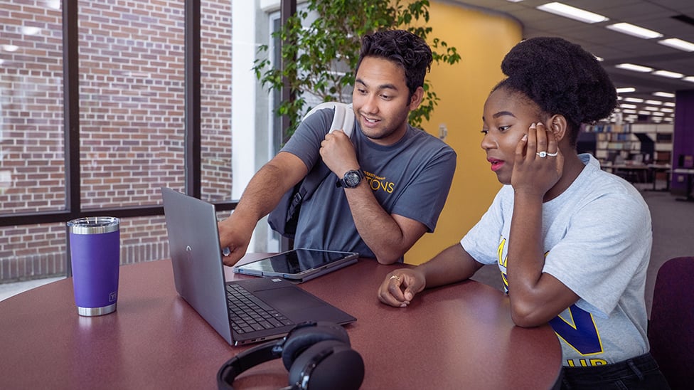 Two students. One sitting at a high top table and the other pointing to something on her computer