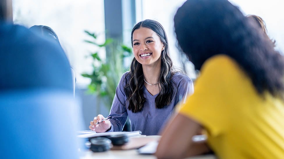 Students around a desk working together, female student smiling.