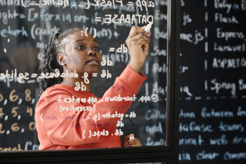 Women writing math equations on a clear board with dry erase markers.