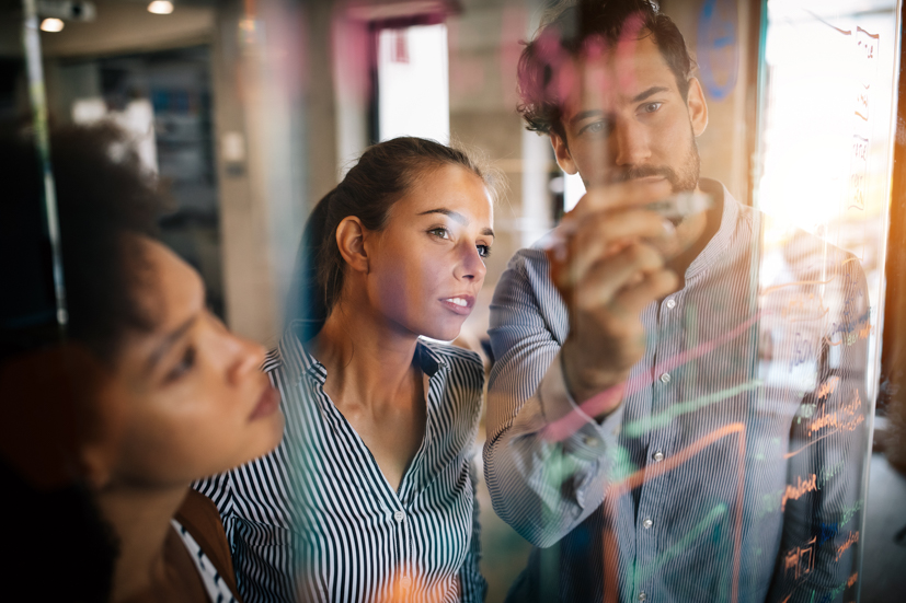 Man writing on clear board and talking to a woman who is looking at the board.