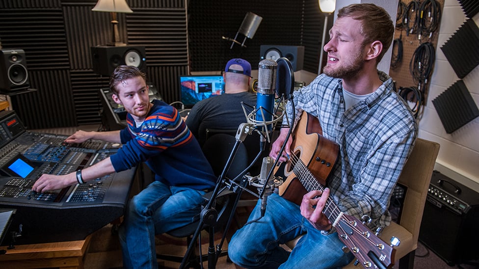 Student playing guitar and singing while other student records song.