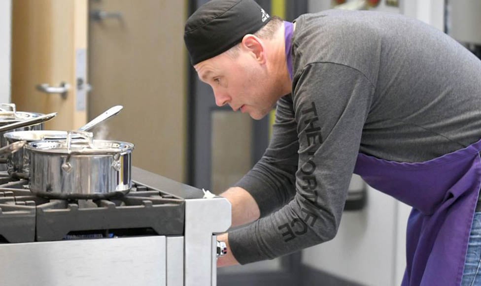 Studet working on the state of the art ovens in the Foods Lab. He is taking something out of the oven while stainless steel cookware is on the top of the stove. 