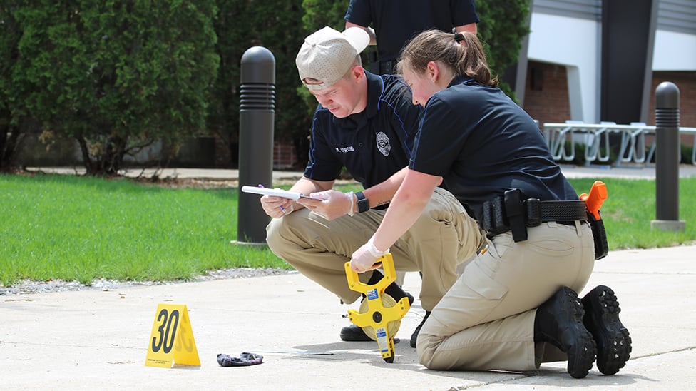 Two students kneeling on the ground looking at paperwork.