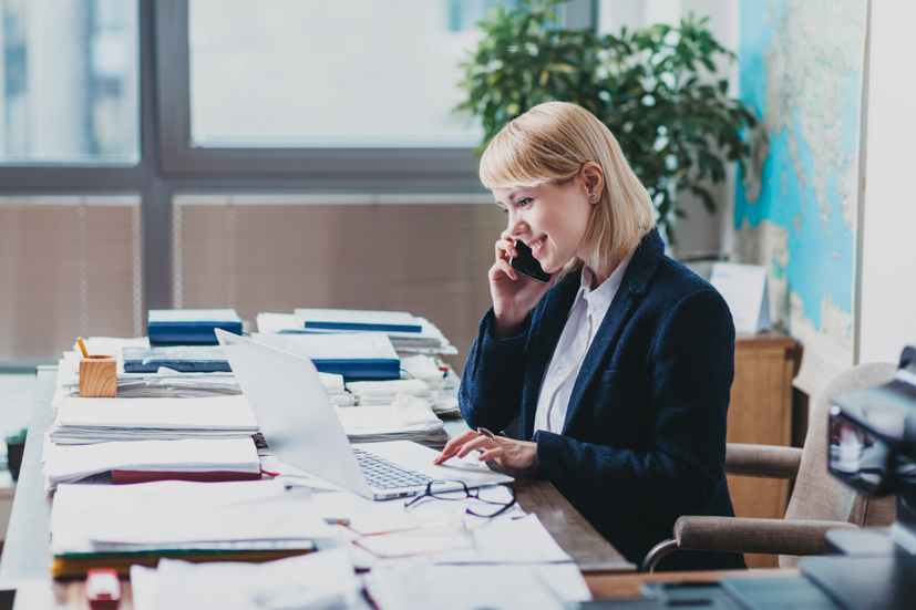 Women sitting at a desk talking on the phone with papers all over the desk.