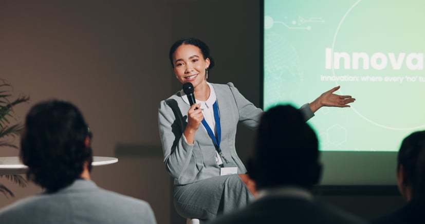 Women giving a speech in a room with the big screen behind her.