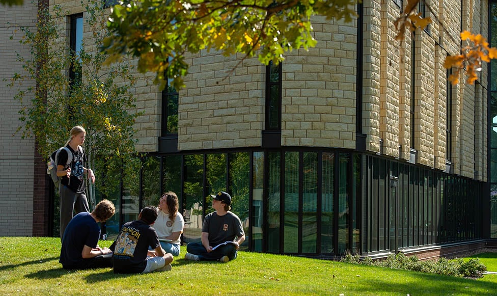 Students sitting outside on the lawn of campus. Sitting in circle talking. It's a beautiful fall day