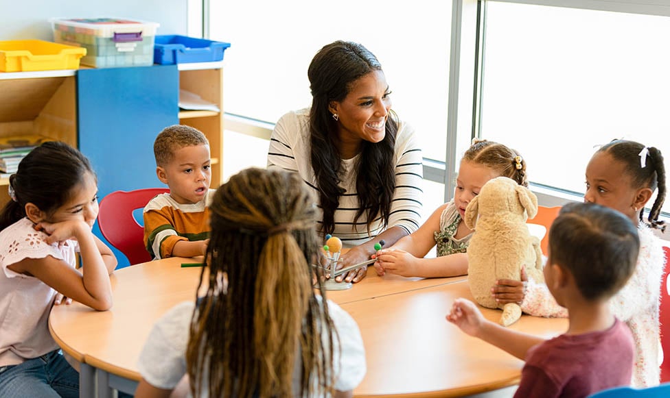 Diverse young teacher sitting at a table with a few elementary students working together and smiling