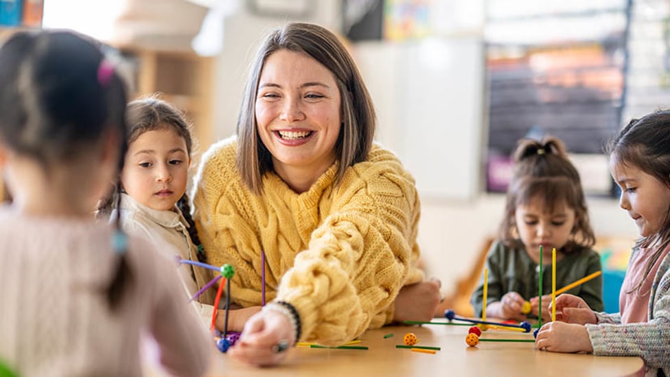 teacher reaching across the table to hand something to a school age child with other children sitting around