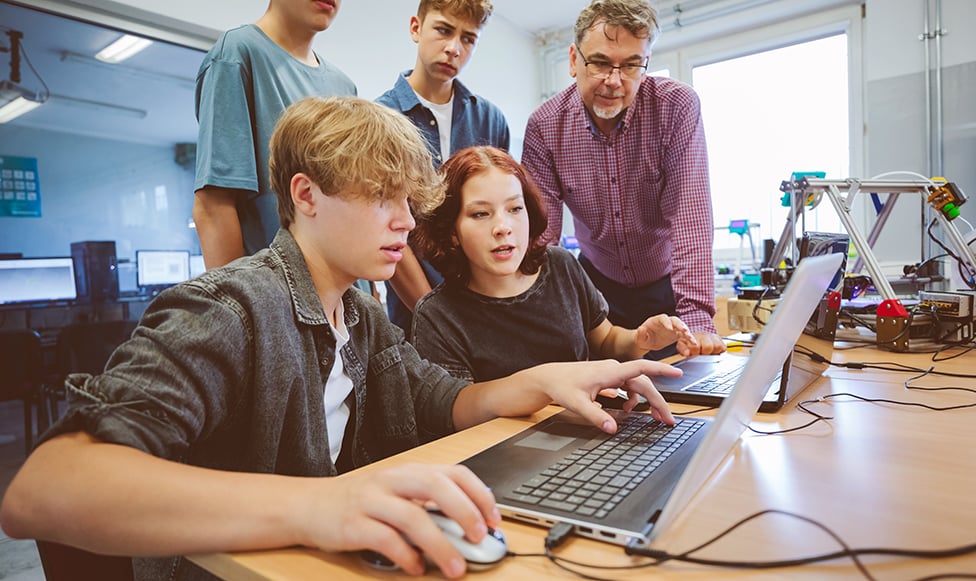 Students working on a computer while teachers look over their shoulders.