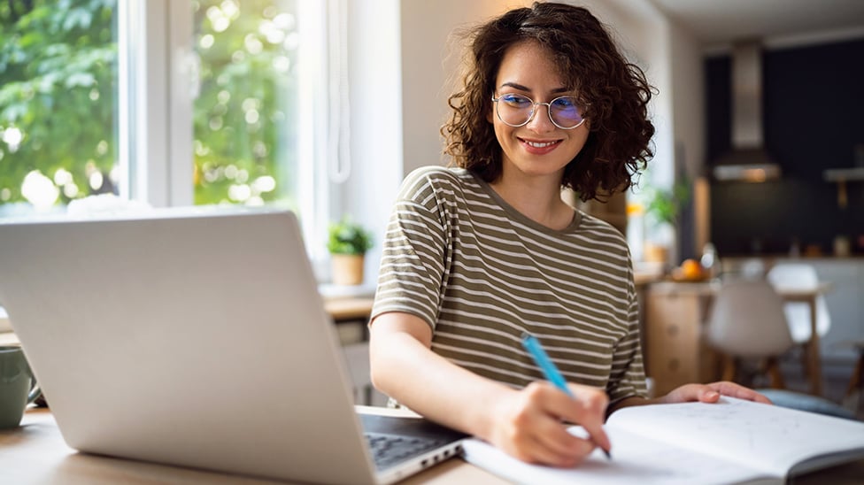 Woman looking at laptop and writing in her notebook.