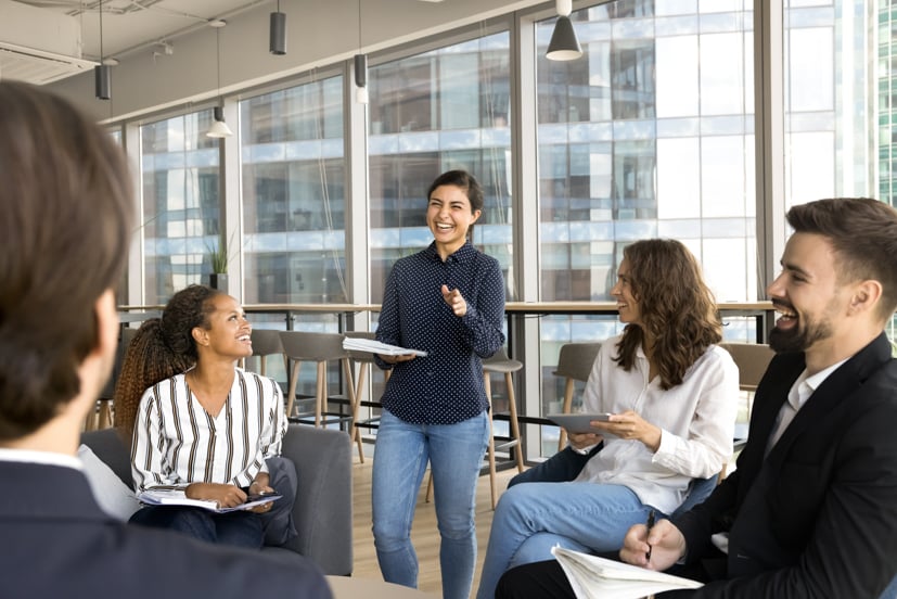Group of men and women in a circle having a discussion, leader of discussion standing and smiling.