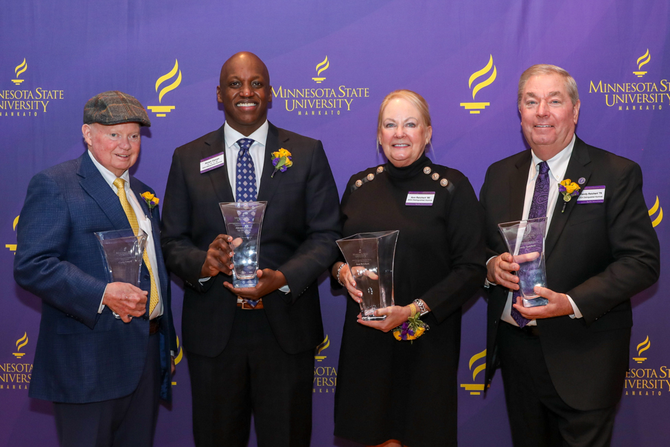 2024 Distinguished Alumni Awards, 4 people standing in front of a MSU backdrop with their glass vase awards.