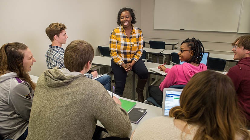 A faculty member leaning on the front desk in a classroom with about 10 students around her listening intently. 