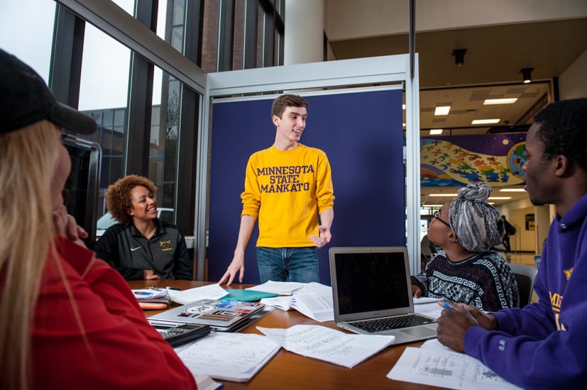 Group of students studying, sitting at a table, a male student standing and talking to them all.