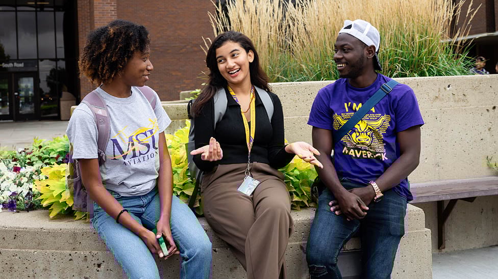 Three international students sit outside on campus with the CSU building behind them.