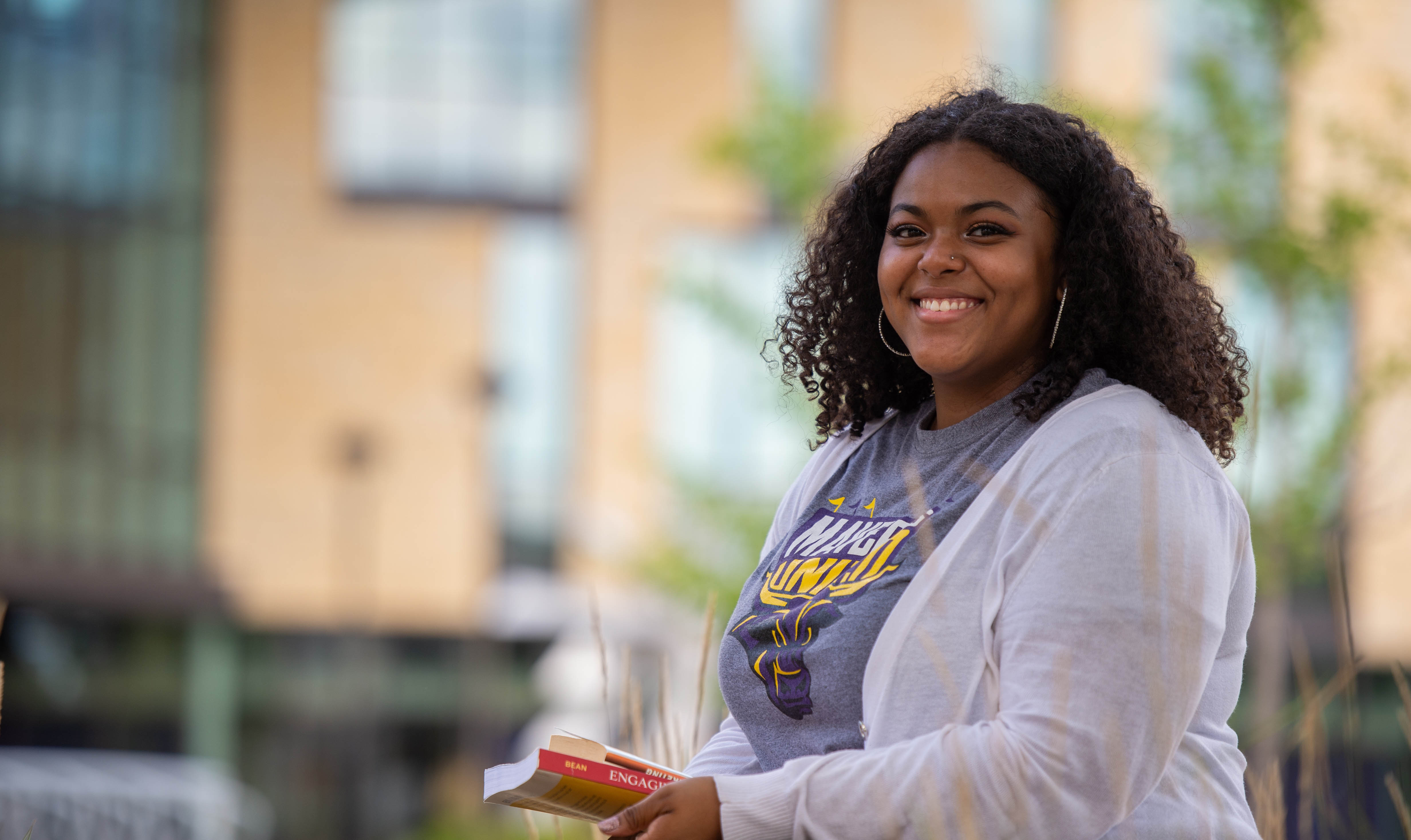 Student sitting on a bench outside on campus holding a book and smiling at the camera