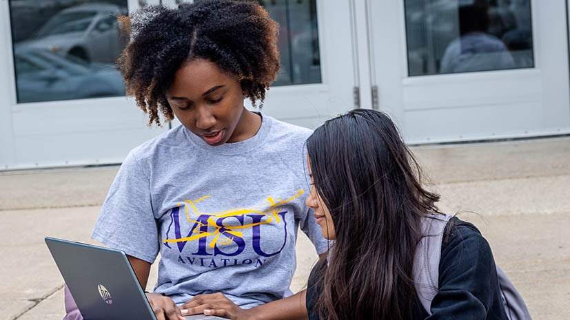 Two international students sitting outside on campus. one is helping the other with some items on the laptop.
