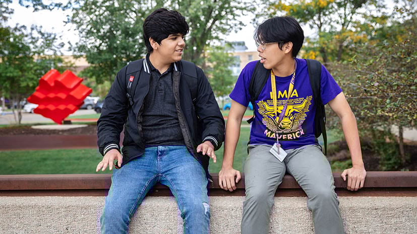 Two students sitting outside on campus in front of some artwork. They are talking to each other.