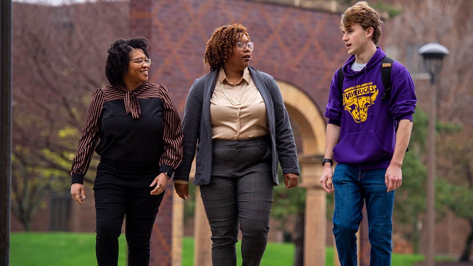 Three people walking on campus. One is speaking and the others listening