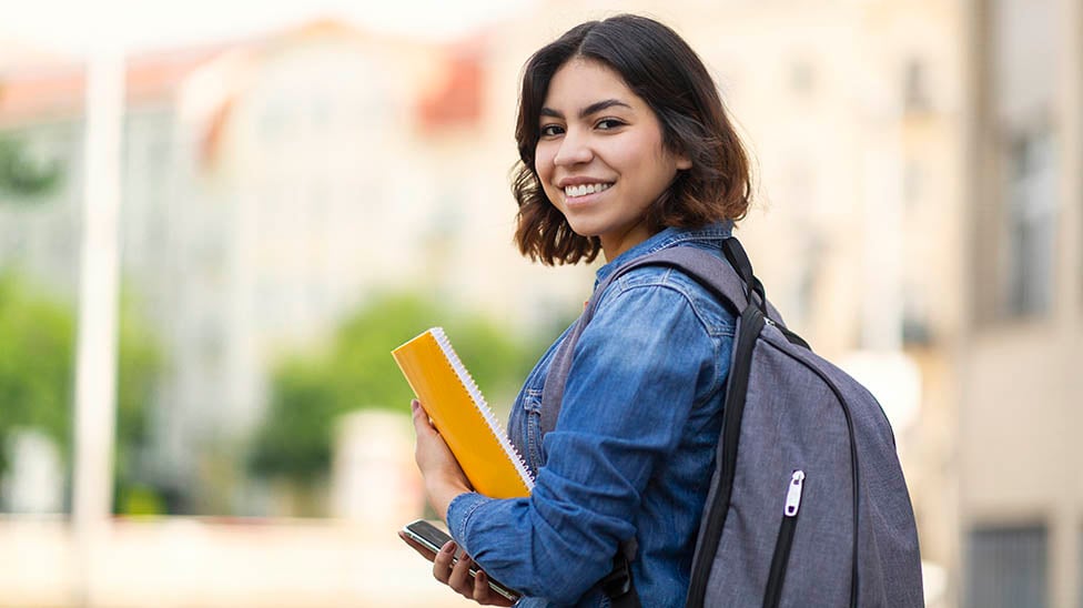 student on campus with backpack and gold folder smiling
