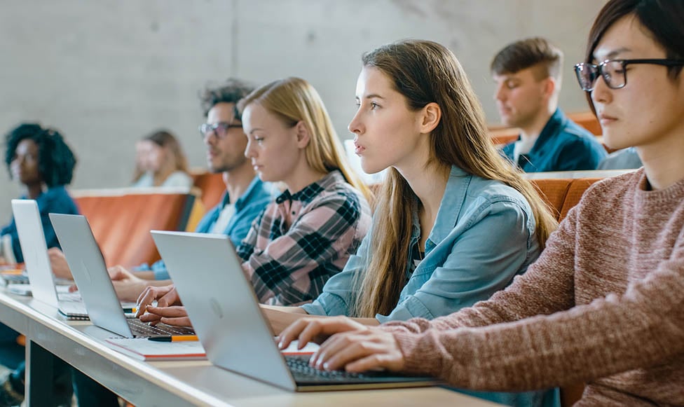 female students in college classroom taking notes on their laptops
