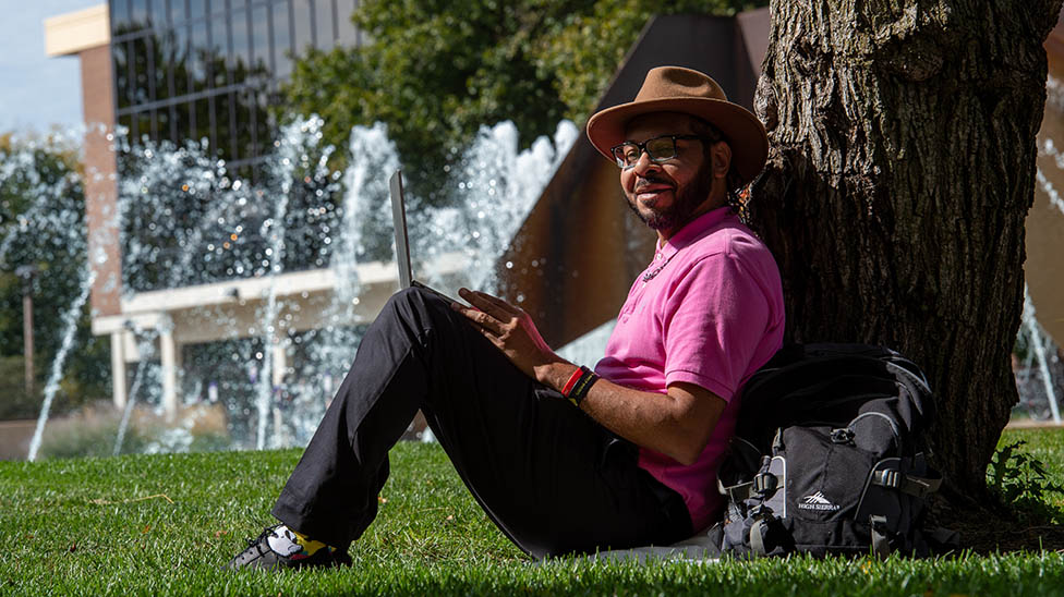 A non-traditional student sits outside on campus against a tree with his backpack and school work. 