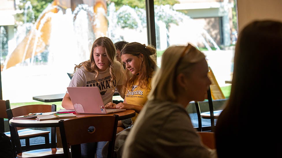 Student female students sitting in the Student Union at a table look at a computer together. The fountain is in the background and other students are sitting at tables nearby. 