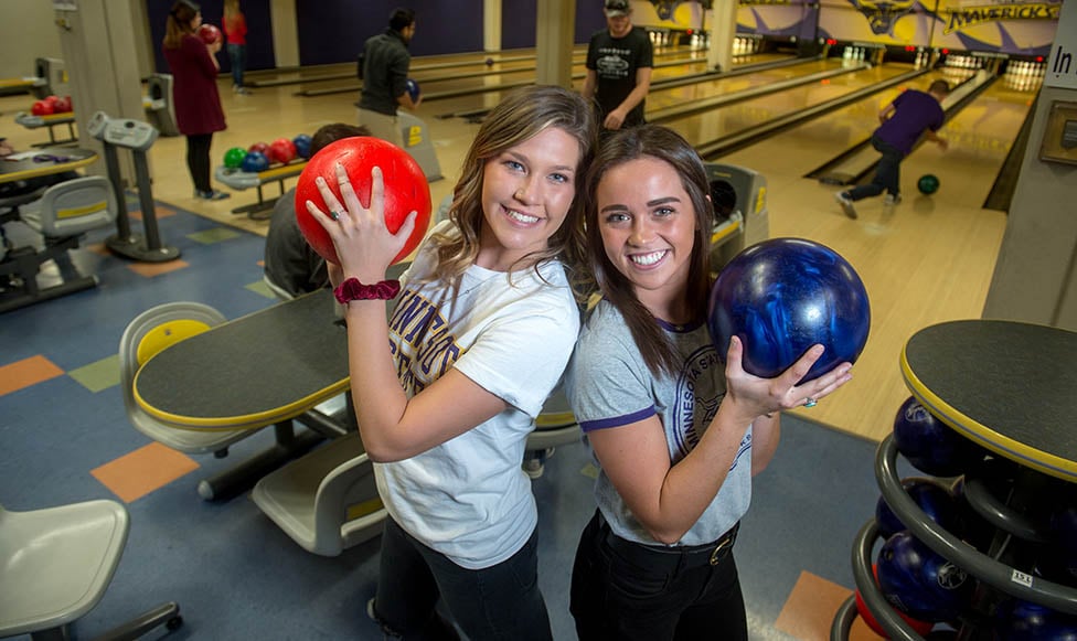 two students posing with bowling balls and the bowling alley behind them