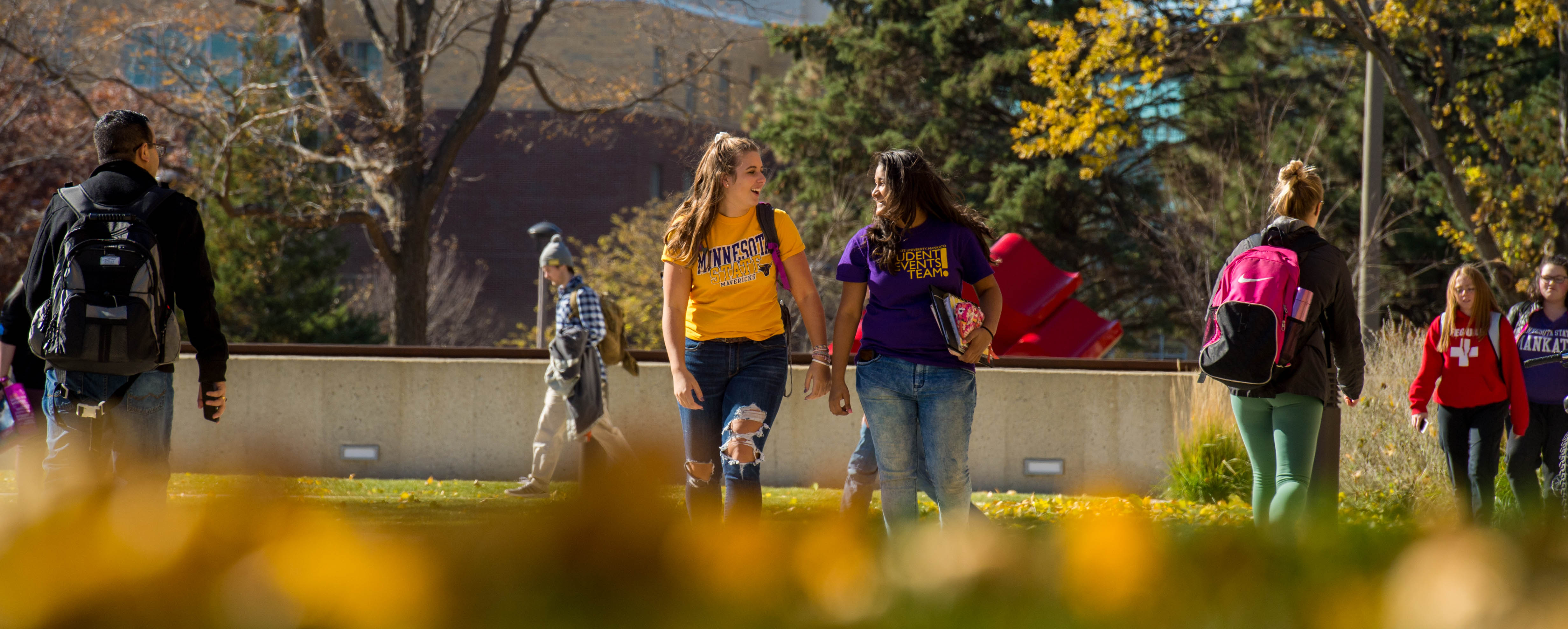 students walking on campus in the fall. Two female students talking and laughing with one another in the forefront