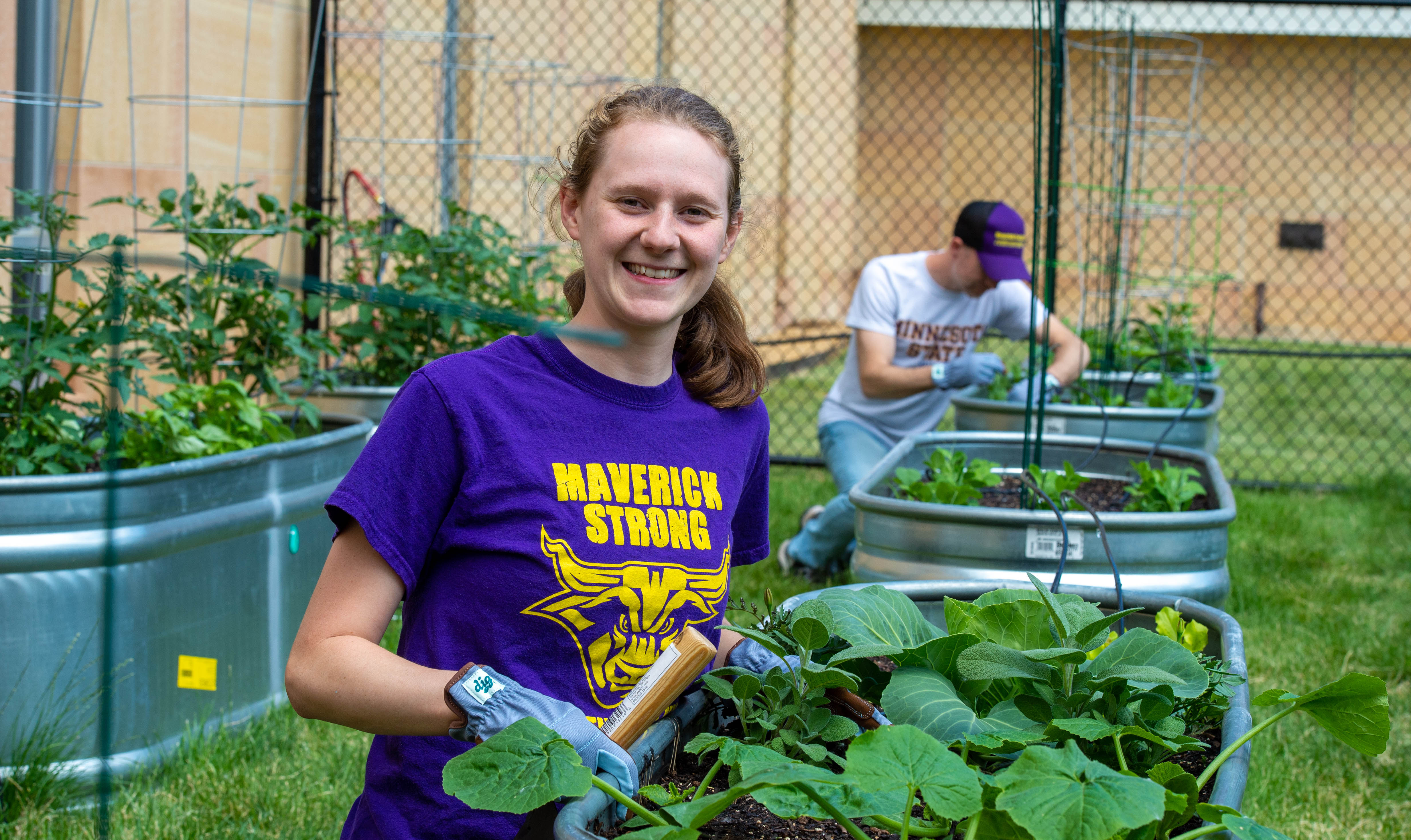 image of female student in the garden smiling with a male student in the background digging in the dirt