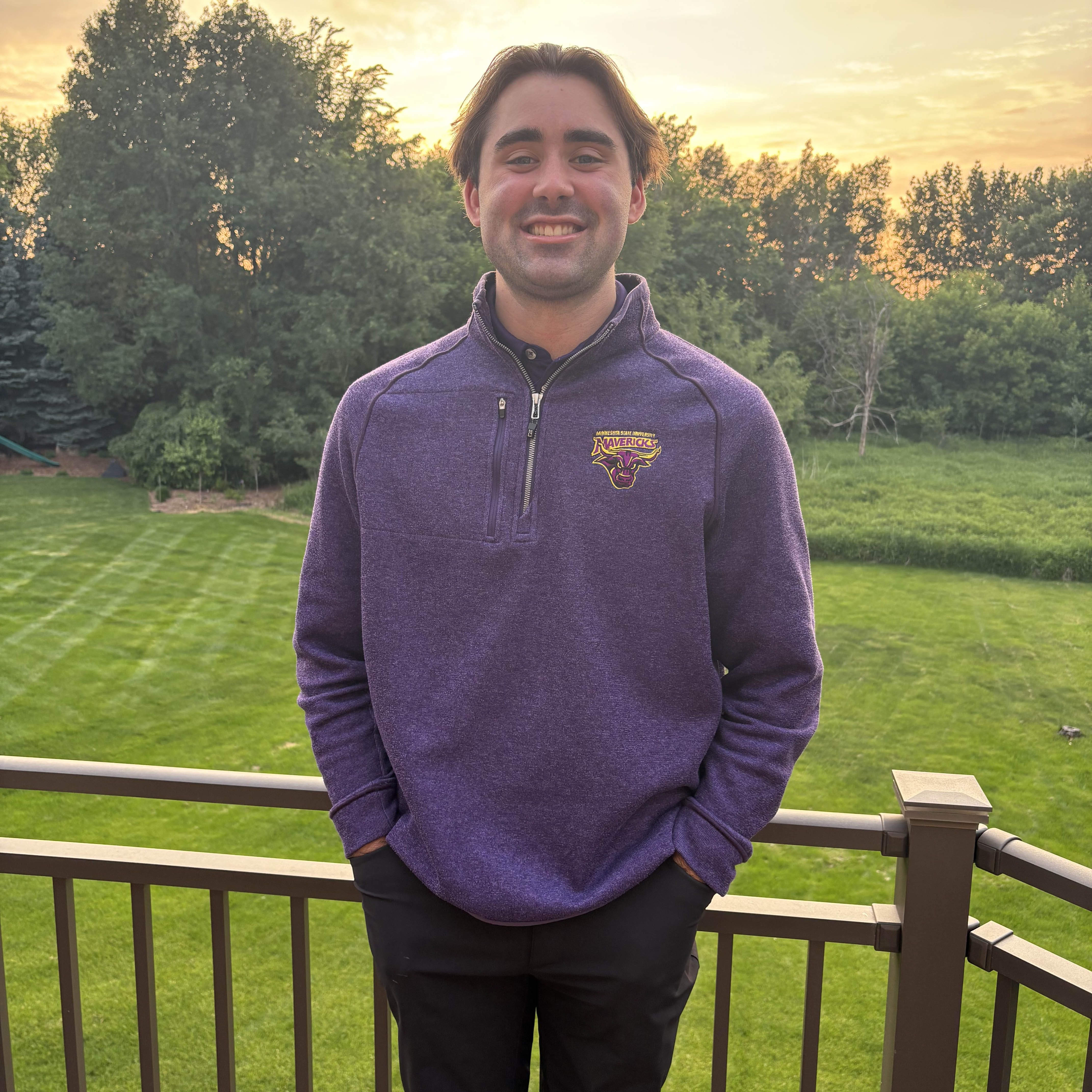  Aiden Hall wearing MSU Maverick gear standing outside on a porch with a sunset in the background. 