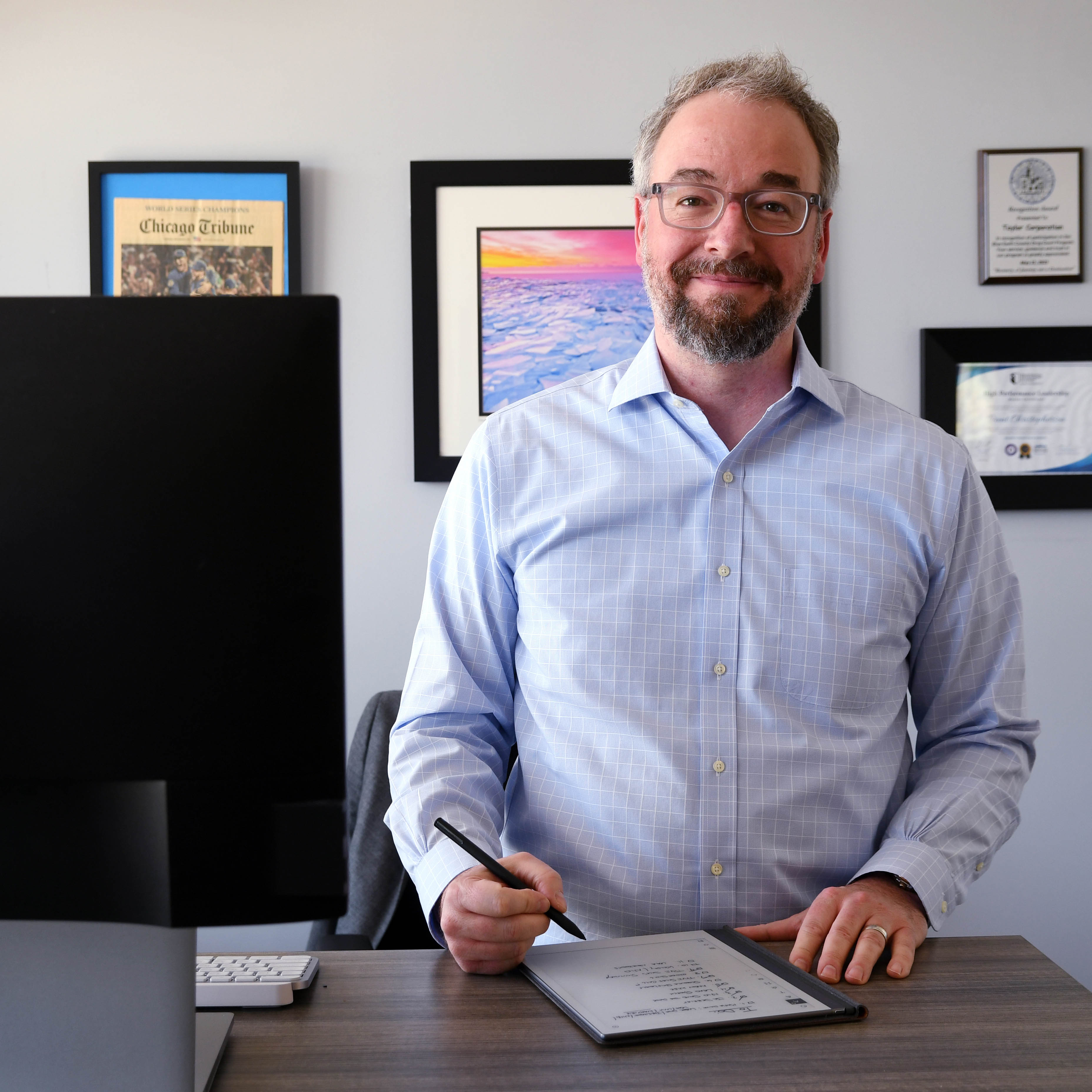 Brent Christopherson posing at his standup desk with a tablet and monitor in front of him. Art work and awards on the wall behind him. 