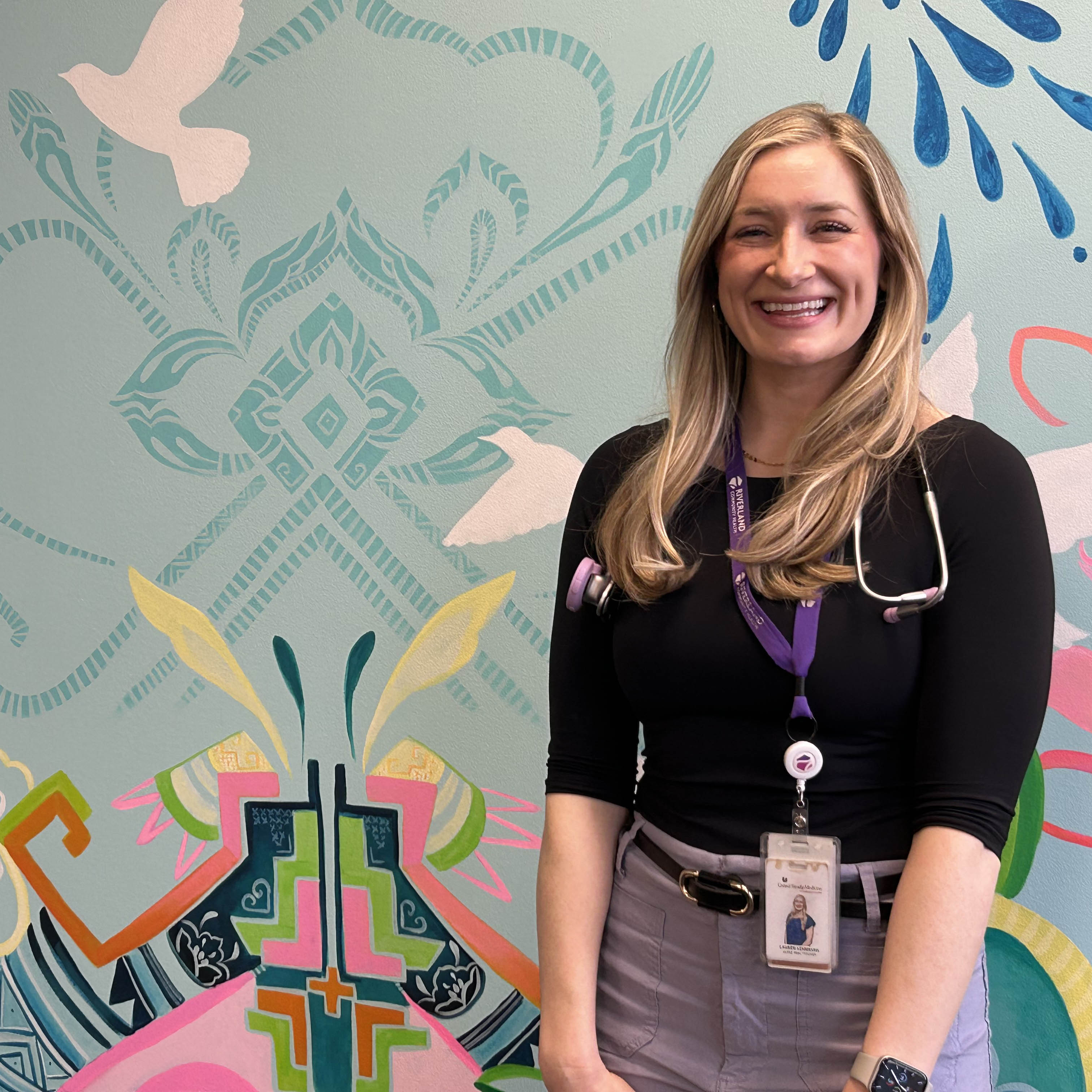 Lauren Lehmkuhl, a nurse practitioner, standing in front of a colorful mural and smiling at the camer. Lemkuhl is wearing a lanyard and stethoscope around her neck