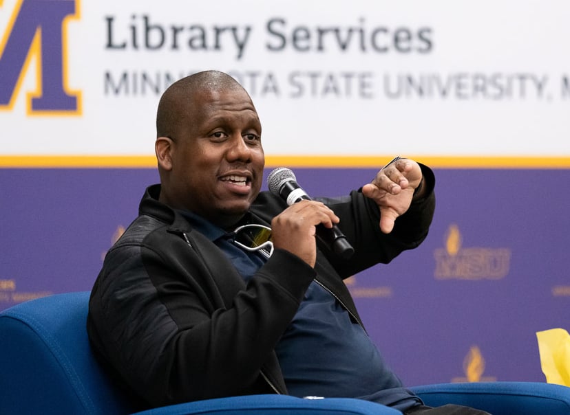 man sitting on stage talking with a microphone and has his hand up