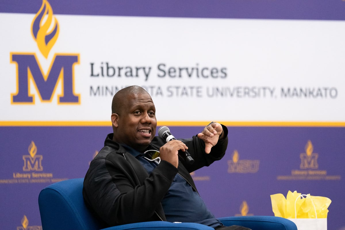 Man sitting on a chair on a stage with a microphone talking to a crowd.