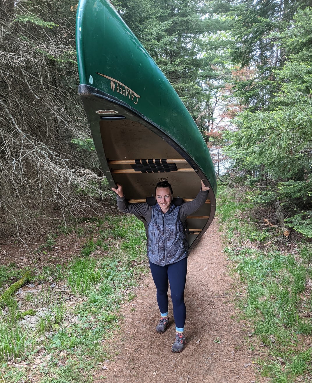 women with dark hair, wearing outdoors attire holding a canoe above her head and walking down a tail in the woods