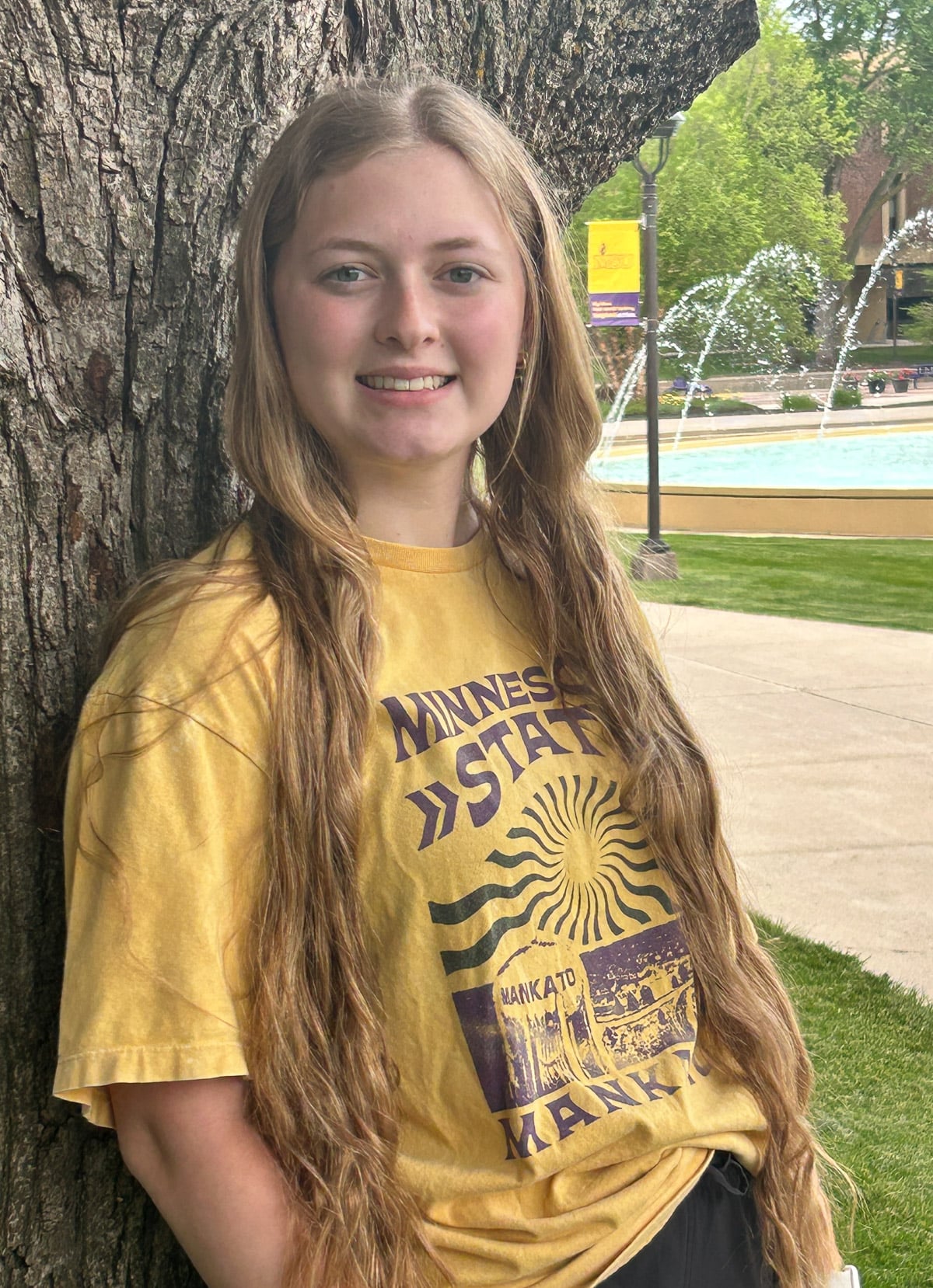 women with light brown hair and wearing a yellow shirt leaning against a tree