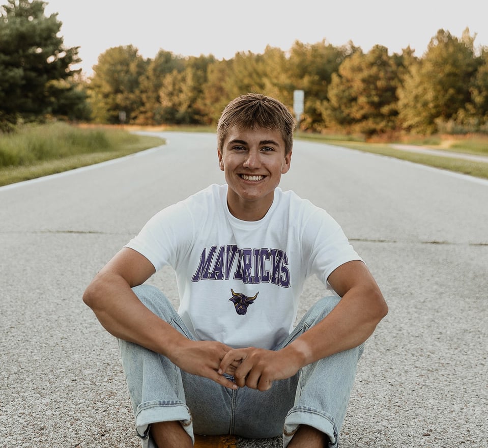 man sitting in the middle of a road on a sunny evening smiling with trees in the distance