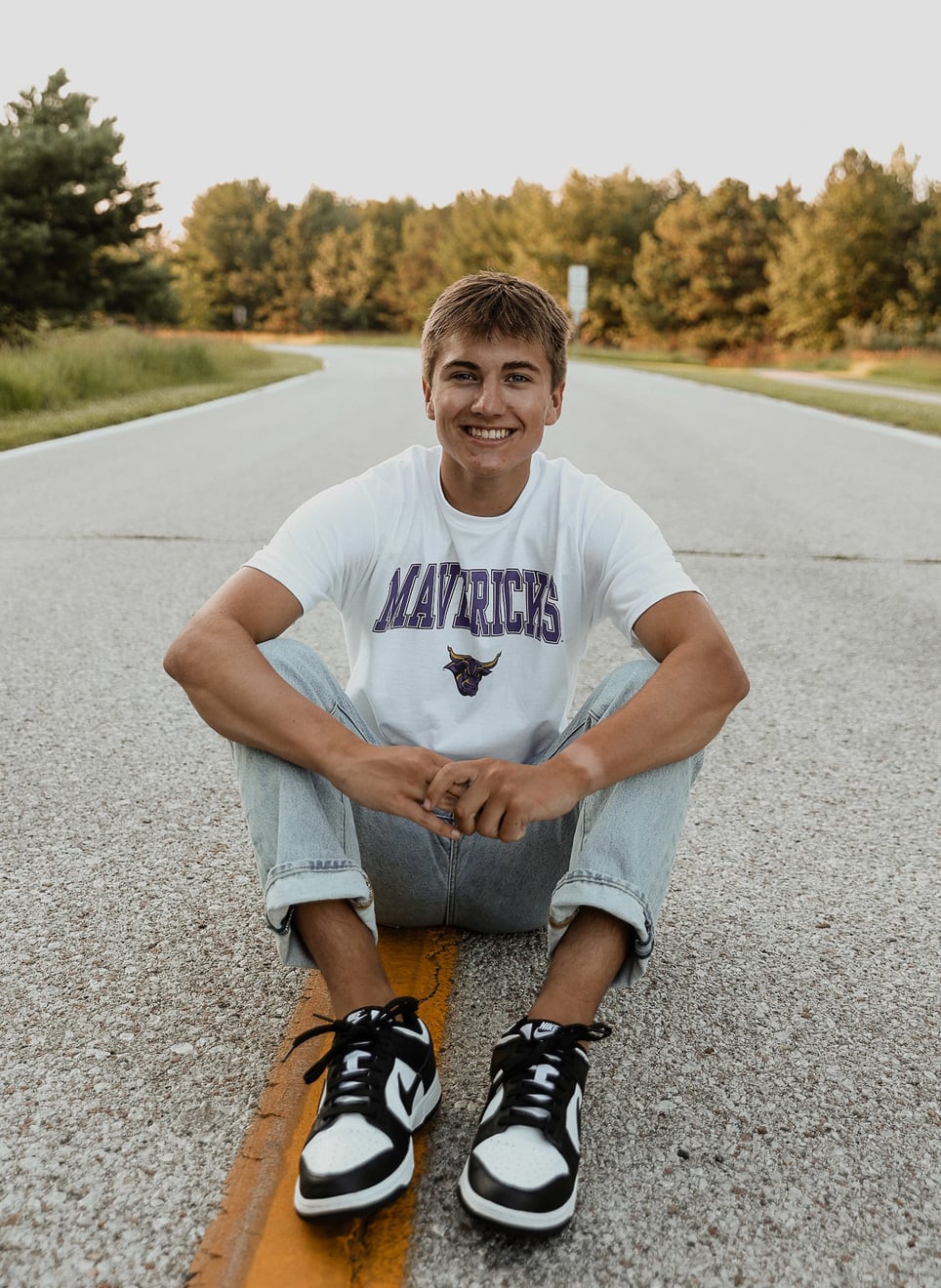 man sitting in the middle of the road cross-legged with a MSU maverick shirt on with trees in the distance