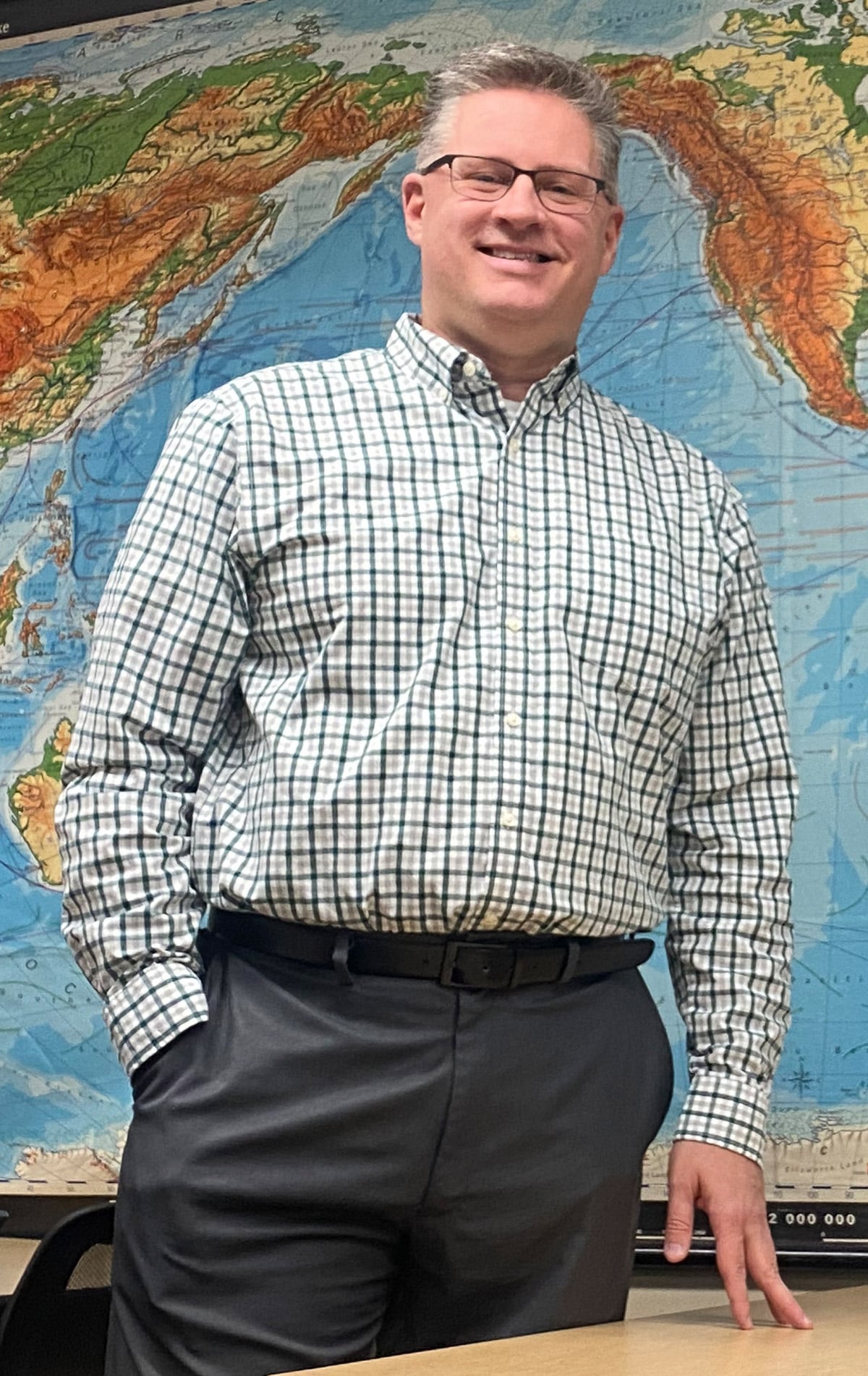man with gray hair and glasses wearing business attire standing in front of a map of the world