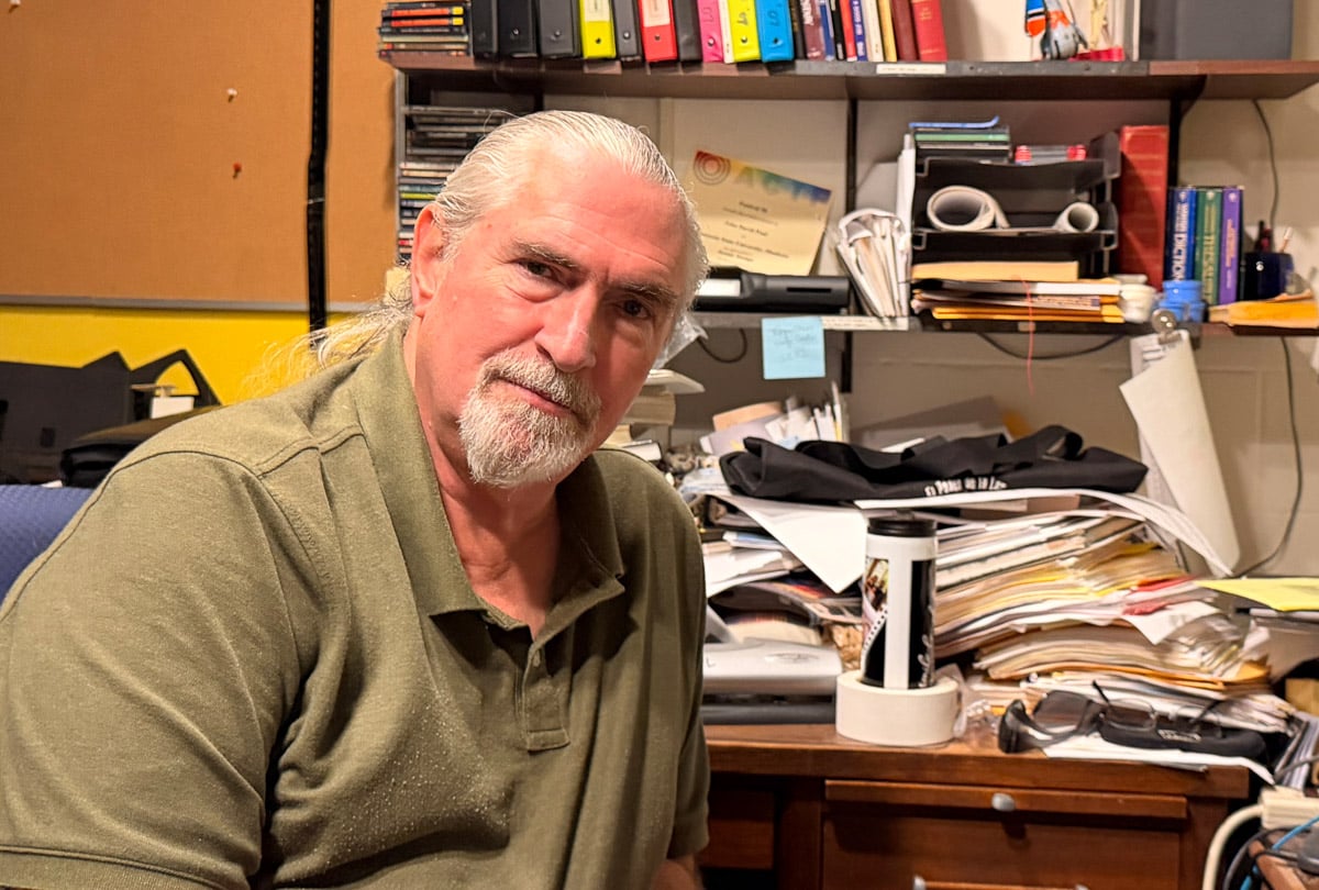 man in a green shirt smiling sitting in front of a desk with many papers and folders on the desk