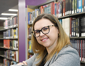 women with glasses leaning on a shelf with bookshelves filled with books in the background