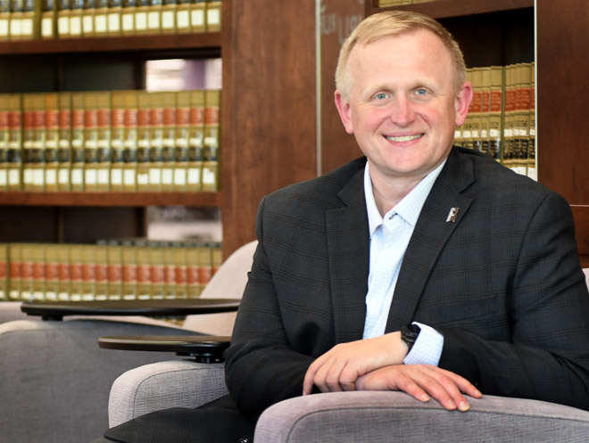 man with blonde hair wearing a black suit sitting a chair in front of a bookcase