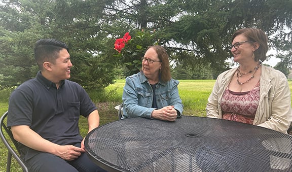 One man and two women sitting around a table outside surrounded by green trees.