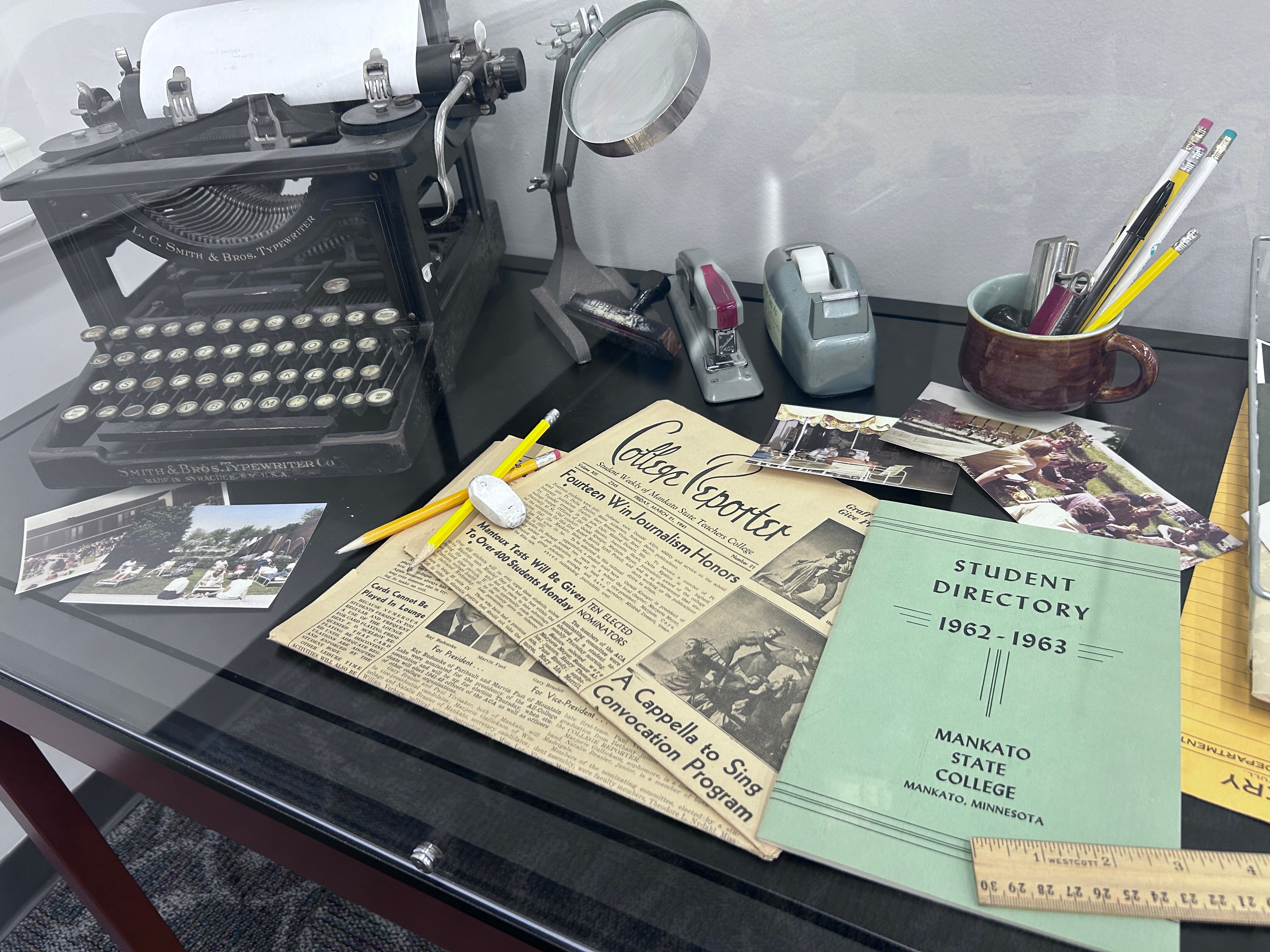 Desk with an old typewriter, newspapers, student directory, rulers, pens and pencils. 