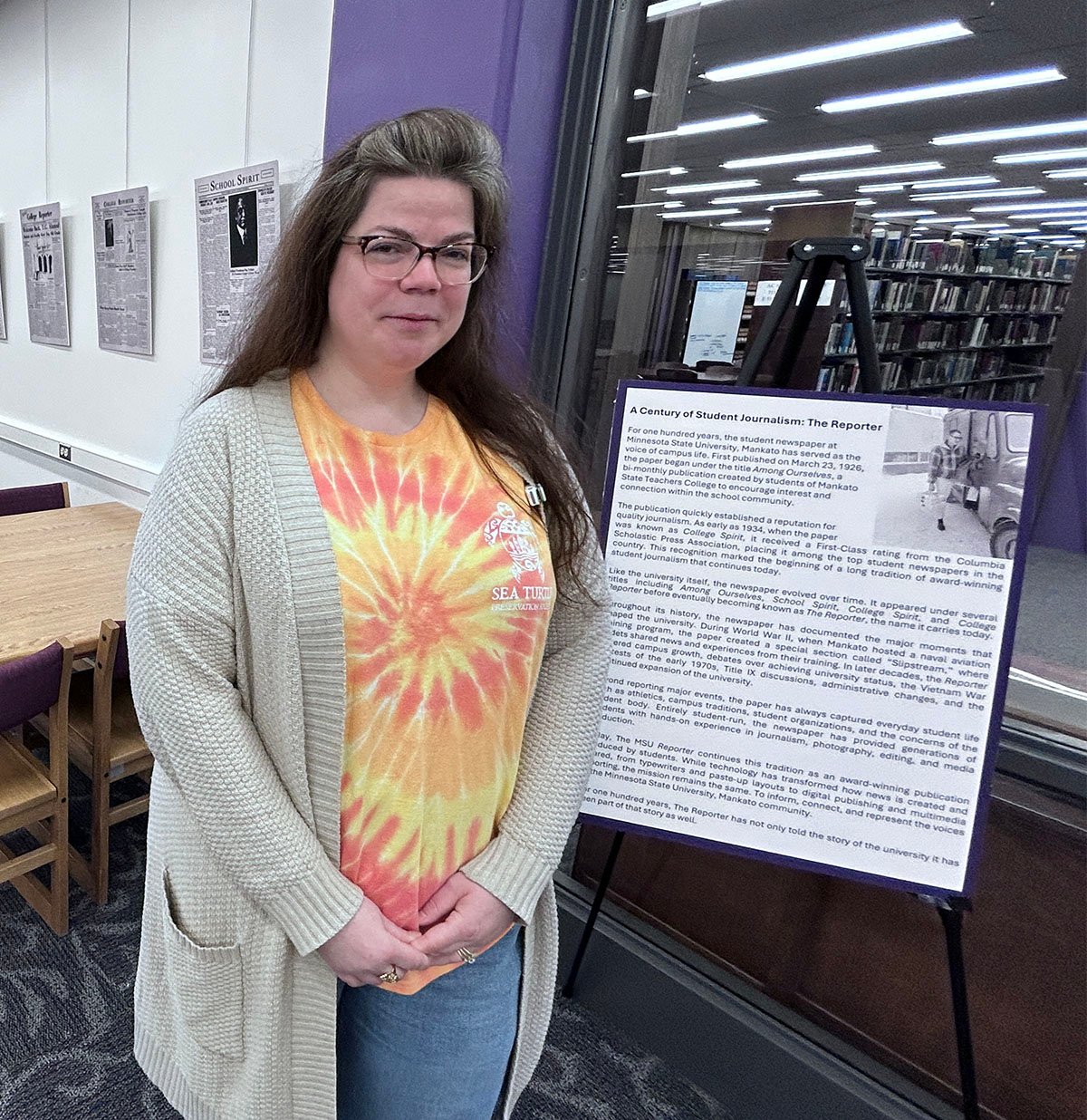 Kelli Flink standing near a poster board about a century of student journalism in the library. 