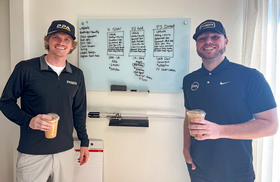 two men with black shirts on and baseball hats holding a coffee standing by a board with notes written on it