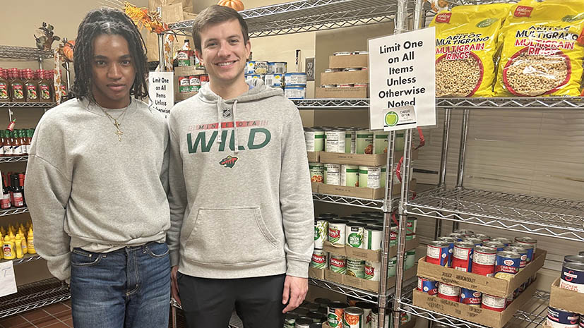 Two students standing in front of the food shelves where students can shop for food. There are bottles of sauce, mustard, soup and more on the shelves. Both students are smiling.