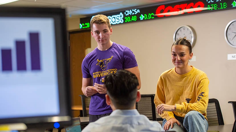 students in the finance lab talking through a work with the stock market ticker in the background