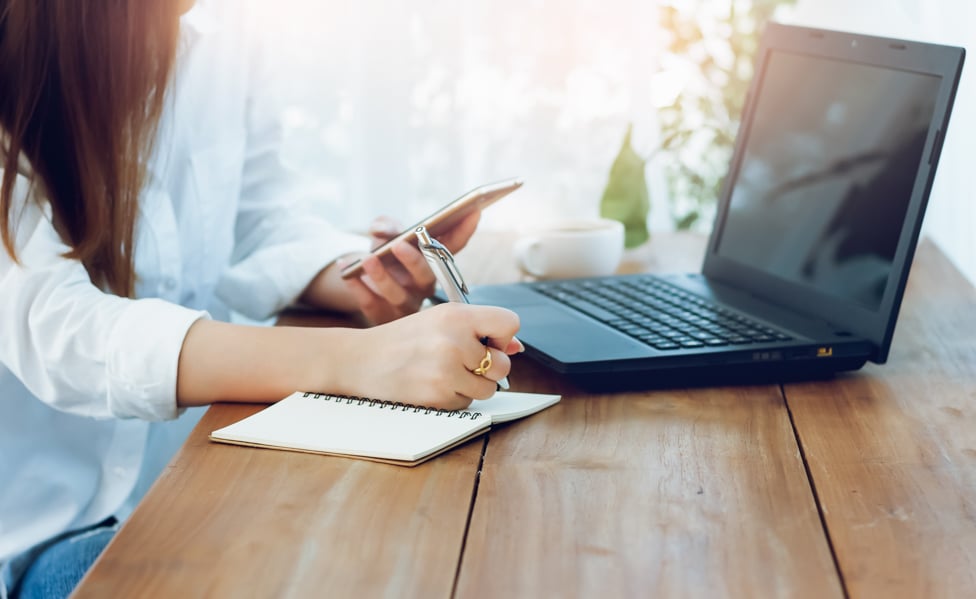 Women sitting in front of computer, holding phone and writing in a notebook.